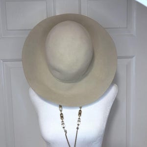 May include: A beige fedora hat with a wide brim. The hat is displayed on a white mannequin head. A long necklace with pearls and decorative beads hangs in front of the mannequin.