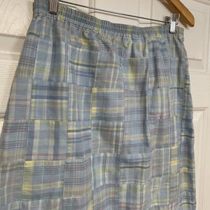 May include: A light blue skirt with a patchwork design. The skirt features a plaid pattern in shades of blue, yellow, and pink. The skirt is hanging on a wooden hanger.
