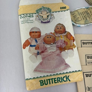 May include: A Butterick sewing pattern envelope, number 6980, for Cabbage Patch Kids Preemie clothes. The front features three dolls wearing a white outfit, a pink dress, and a yellow dress, with the text Cabbage Patch Kids Preemie and Butterick.