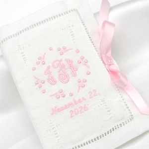 May include: A white rectangular fabric pouch featuring pink embroidered monogram initials surrounded by a floral wreath design. Below the monogram, the text reads November 22, 2026. A light pink satin ribbon is tied in a bow on the right side.