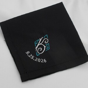 May include: A square black fabric handkerchief featuring an embroidered design in the center. The embroidery consists of a white swirling letter K surrounded by teal geometric accents, with the date 9.25.2026 stitched in white below it.