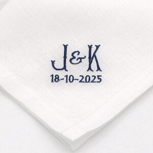 May include: White linen handkerchief with embroidered navy blue initials "J & K" and the date "18-10-2025". The handkerchief is folded, showing a corner with the embroidered details.