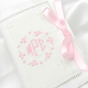 May include: A white rectangular linen handkerchief featuring a delicate hemstitched border and a pink embroidered monogram with the letters MPE surrounded by a small floral wreath. A light pink satin ribbon is tied around the corner of the fabric.