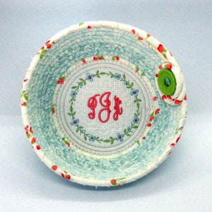 May include: A white fabric bowl with a blue and white floral pattern. The bowl has a round, embroidered center with the initials "J.B." in pink. The bowl has a green button on the side.