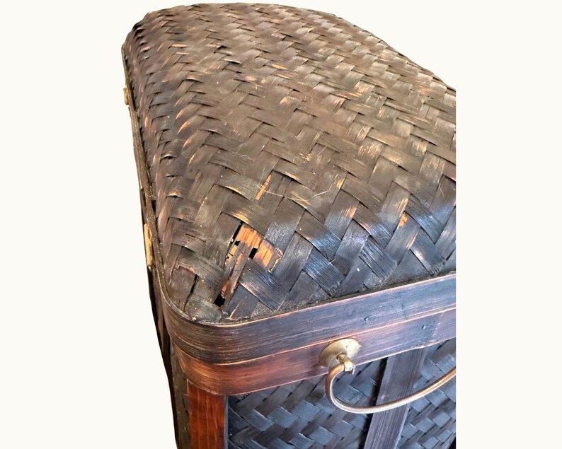 May include: A dark brown woven trunk with a curved top. The trunk has a rectangular shape with a dark brown wooden frame and a metal handle. The woven material has a textured appearance with visible wear and tear.