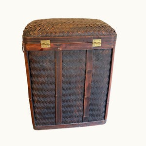 May include: A rectangular, dark brown woven basket with a hinged lid. The basket features a tightly woven pattern and is secured with brass-colored metal hinges and clasps. Vertical wooden supports and a handle are visible.
