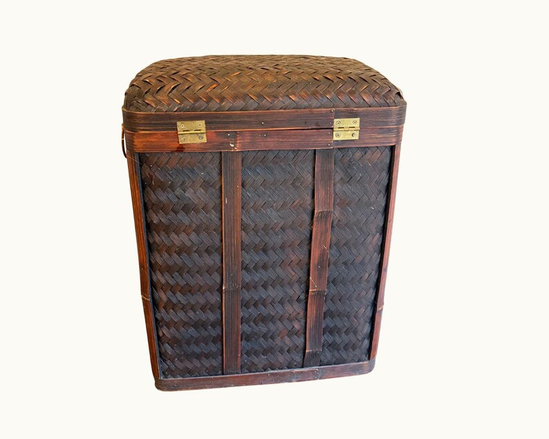 May include: A rectangular, dark brown woven basket with a hinged lid. The basket features a tightly woven pattern and is secured with brass-colored metal hinges and clasps. Vertical wooden supports and a handle are visible.