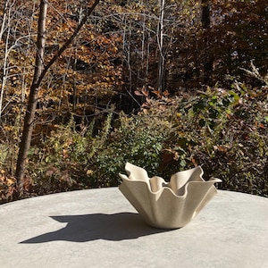 May include: A white ceramic bowl with a ruffled edge, sitting on a gray table. The bowl is in the shape of a flower, with five petals.
