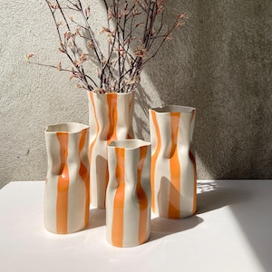May include: Four ceramic vases with a white and orange striped pattern. The vases are of different sizes and are arranged on a white surface.