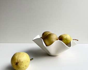 May include: A white ceramic bowl with a wavy edge, filled with four green pears. The bowl is sitting on a white surface.