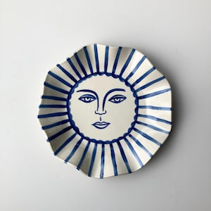 May include: A ceramic plate with a sun face design. The plate has a white background with a blue sun face in the centre and blue radiating stripes around the edge. The plate has a wavy, scalloped rim.