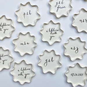 Könnte beinhalten: Zehn weiße Keramikteller mit gewellten Rändern, jeder mit einer anderen handschriftlichen Botschaft in schwarzer Tinte. Die Botschaften beinhalten Namen und Sätze wie "a lifetime with you" und "g & b forever".