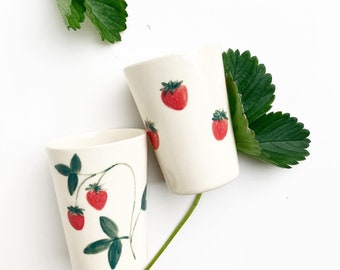 May include: Two white ceramic mugs with strawberry designs. One mug has a single strawberry design, while the other has a strawberry vine design with multiple strawberries.
