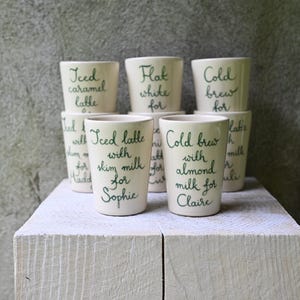 May include: A set of six off-white ceramic cups arranged in two rows. Each cup features dark green handwritten text specifying various coffee orders, including Iced caramel latte, Flat white, Cold brew, and personalised names like Sophie and Claire.