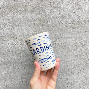 May include: White ceramic mug with a blue and white design of fish and waves. The word "SARDINIA" is written in blue on the mug.