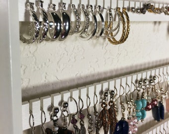 May include: A white wooden earring holder with multiple rows of hooks. The top row is filled with silver hoop earrings, while the bottom row features a variety of dangle earrings with silver, turquoise, and brown accents.