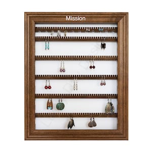 Op de afbeelding: Een houten oorbellenhouder met het woord "Mission" erop gedrukt. De houder heeft vijf rijen haken voor oorbellen. De houder is gevuld met verschillende oorbellen.