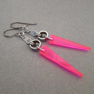 May include: A pair of neon pink, triangular earrings. The earrings have a silver-colored hook and ring design. The pink triangles are approximately 1.5 inches long and taper to a point. The earrings are displayed against a gray background.