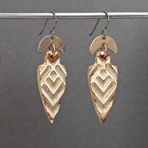 Könnte beinhalten: Ein Paar handgefertigte Ohrringe mit pfeilförmigen Anhängern in Creme mit geometrischem Muster. Die Ohrringe haben einen silberfarbenen Halbmond-Akzent und einen Hakenverschluss. Die Ohrringe werden vor grauem Hintergrund präsentiert, ca. 5 cm lang.