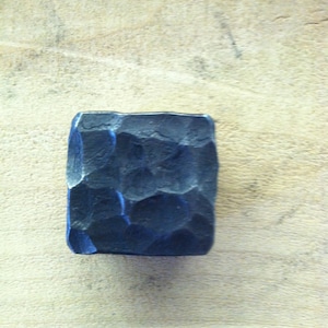 May include: A square, dark blue metal object with a textured, hammered surface. The object is set against a light brown wooden background. The square shape and textured finish suggest a handcrafted item.