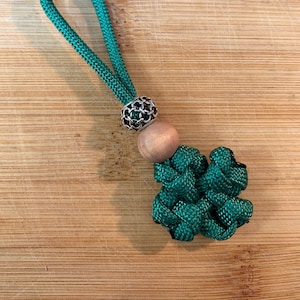 May include: A green paracord keychain with a wooden bead and a silver metal bead. The keychain is tied in a knot that resembles a four-leaf clover.