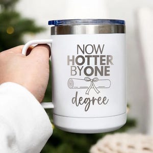 May include: White travel mug with a black and white graphic that says "Now hotter by one degree" and a drawing of a diploma scroll with a ribbon.