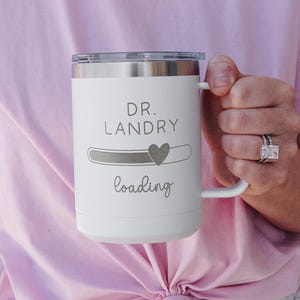 May include: White travel mug with a black handle and a gray inscription that reads "DR. LANDRY loading" with a small heart icon.