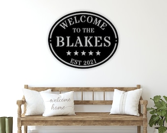 May include: Black oval metal sign with white text that reads "Welcome to the Blakes Est 2021" with five white stars between the words "Blakes" and "Est". The sign is hanging on a white wall above a wooden bench with white pillows.