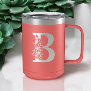 May include: Coral pink stainless steel travel mug with a silver lid and handle. The mug has a white floral design with the letter "B" in the center.