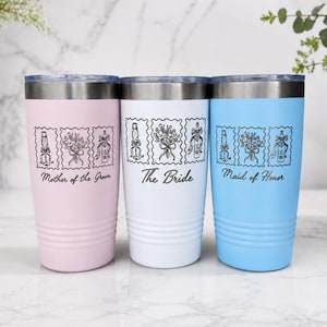 May include: Three stainless steel tumblers in light pink, white, and light blue, each featuring a silver rim and a black line-art graphic of a champagne bottle, bouquet, and gift. The text reads Mother of the Groom, The Bride, and Maid of Honor.