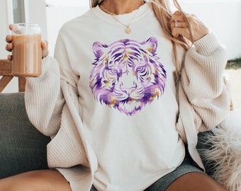 May include: A white sweatshirt with a purple and gold tiger face graphic. The model is wearing the sweatshirt with a white cardigan and sitting on a fluffy white chair.