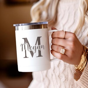 May include: White travel mug with a silver rim and a clear lid. The mug features a large gray letter "M" with the name "Megan" below it. The mug has a white handle.