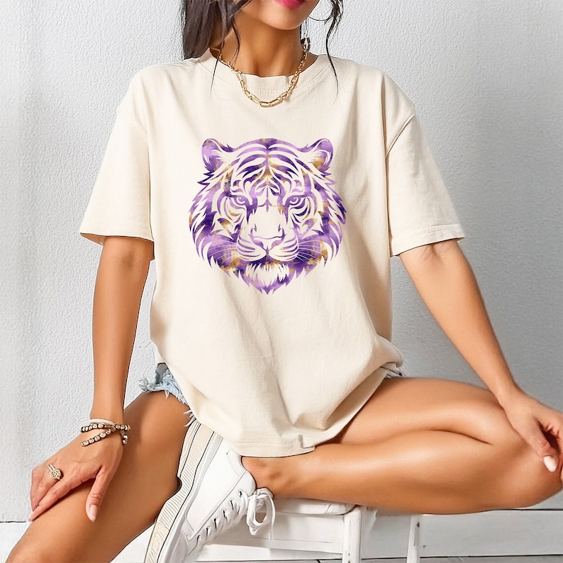 May include: A white t-shirt featuring a purple and gold tiger face graphic. The tiger has a fierce expression and is surrounded by a purple and gold swirl pattern.