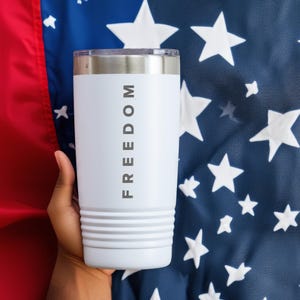 May include: White tumbler with the word "FREEDOM" printed vertically. The tumbler has a silver rim and a ridged design near the bottom. The background features a blue fabric with white stars and a red stripe.