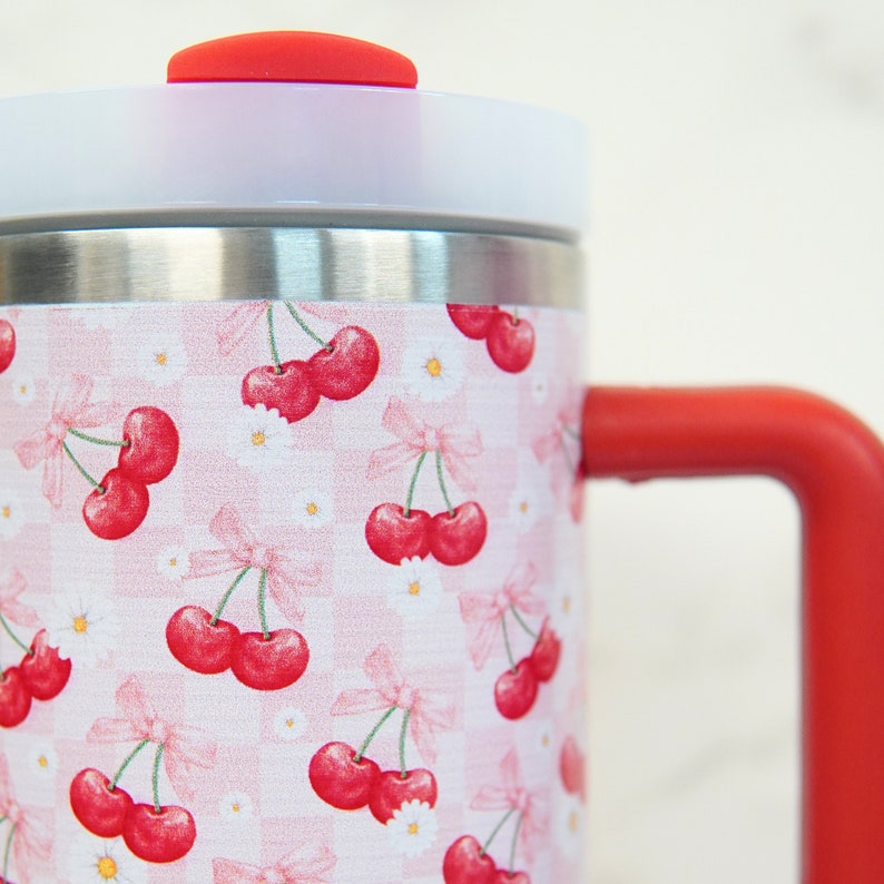 Puede incluir: Un termo de viaje de acero inoxidable con asa y detalles de tapa rojos. El termo presenta un patr&oacute;n repetido de cerezas rojas, lazos rosas y margaritas blancas sobre un fondo a cuadros rosa y blanco.