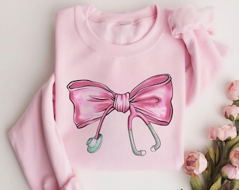 May include: Pink sweatshirt with a pink bow and a stethoscope design. The bow is tied with a gray ribbon.