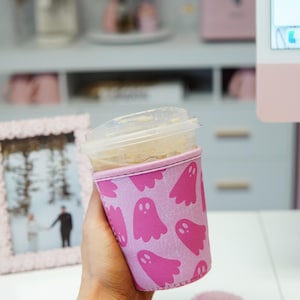 May include: A pink coffee cup sleeve featuring a repeating pattern of pink ghost illustrations. The sleeve is around a clear plastic cup with a lid. A pink flower-shaped coaster is in the foreground.