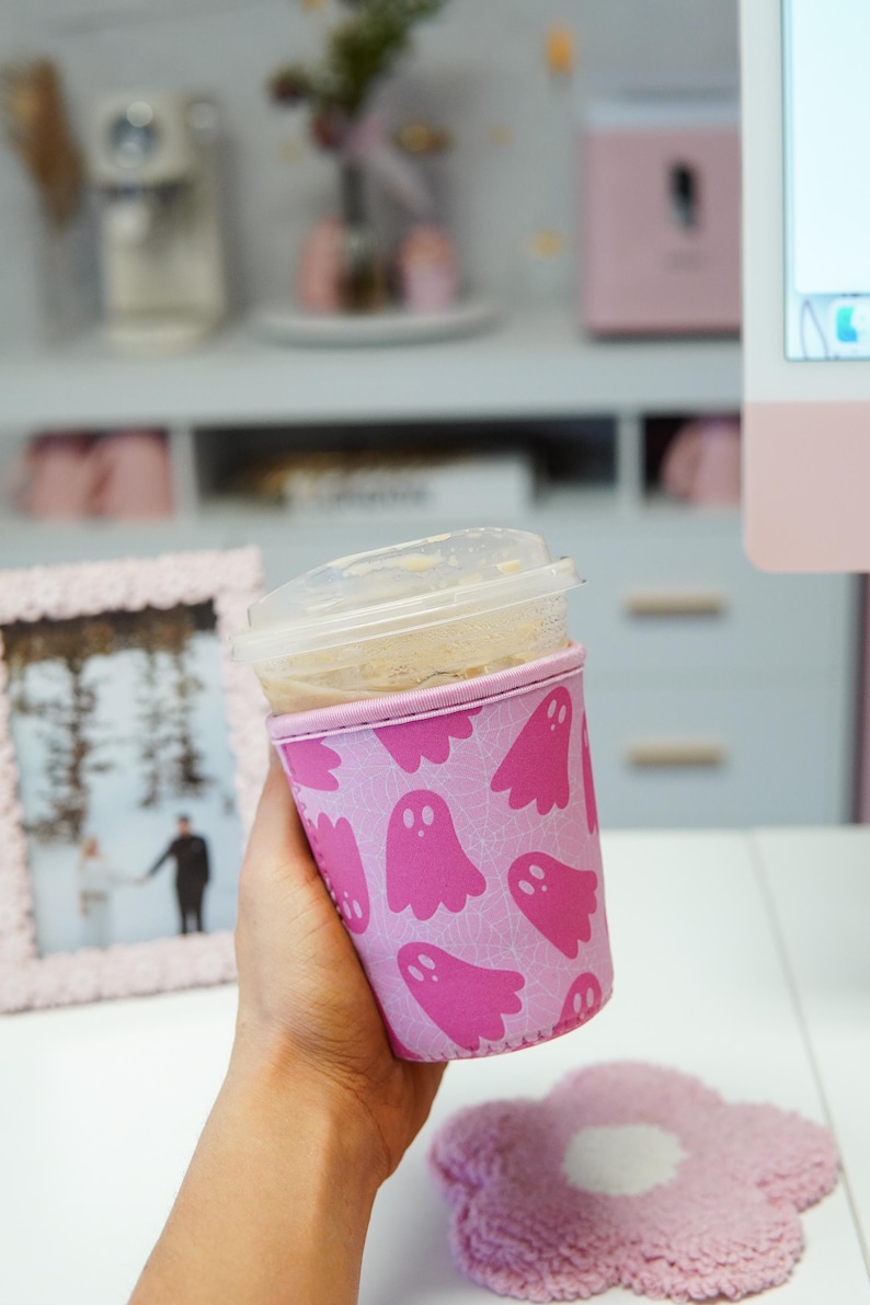 May include: A pink coffee cup sleeve featuring a repeating pattern of pink ghost illustrations. The sleeve is around a clear plastic cup with a lid. A pink flower-shaped coaster is in the foreground.
