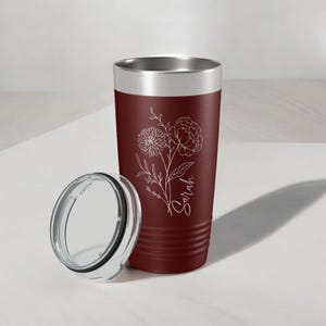 May include: A maroon stainless steel tumbler with a white floral design and the name "Sarah" engraved on the side. The tumbler has a clear lid.