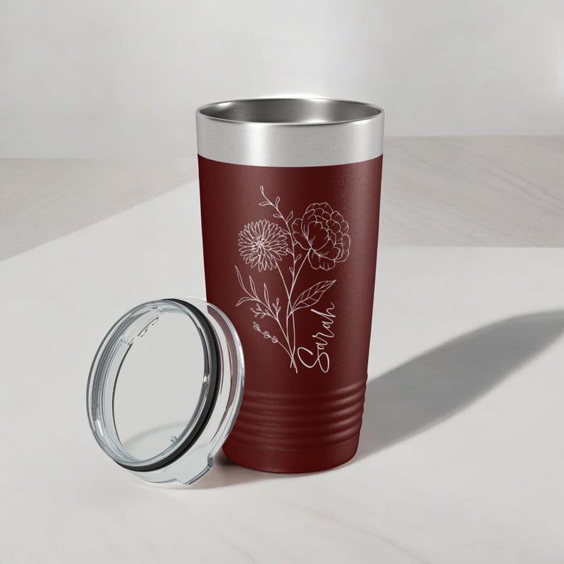 May include: A maroon stainless steel tumbler with a white floral design and the name "Sarah" engraved on the side. The tumbler has a clear lid.