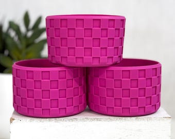 May include: Three fuchsia-colored silicone water bottle sleeves stacked on a white surface. Each sleeve has a woven basketweave pattern. The sleeves are cylindrical and designed to fit around a water bottle.