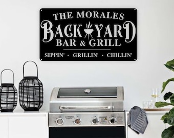 May include: Black metal sign with white text that reads "The Morales Backyard Bar & Grill Sippin' Grillin' Chillin'" with a grill icon. The sign is hanging on a white wall above a black and silver gas grill.