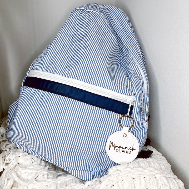Pu&ograve; includere: Zaino a righe blu e bianche con cerniera bianca e una striscia di accento blu navy. Lo zaino presenta un'etichetta rotonda bianca con il testo "Maverick Dupuis" e un portachiavi dorato.