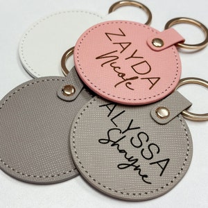 Pu&ograve; includere: Una collezione di portachiavi personalizzati in vari colori. I portachiavi sono rotondi con una superficie strutturata e presentano nomi in un carattere scritto a mano. I portachiavi hanno portachiavi e chiusure color oro.