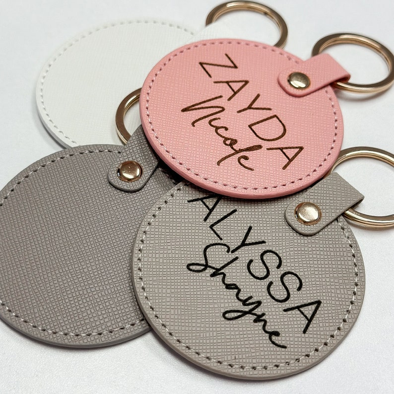 Pu&ograve; includere: Una collezione di portachiavi personalizzati in vari colori. I portachiavi sono rotondi con una superficie strutturata e presentano nomi in un carattere scritto a mano. I portachiavi hanno portachiavi e chiusure color oro.
