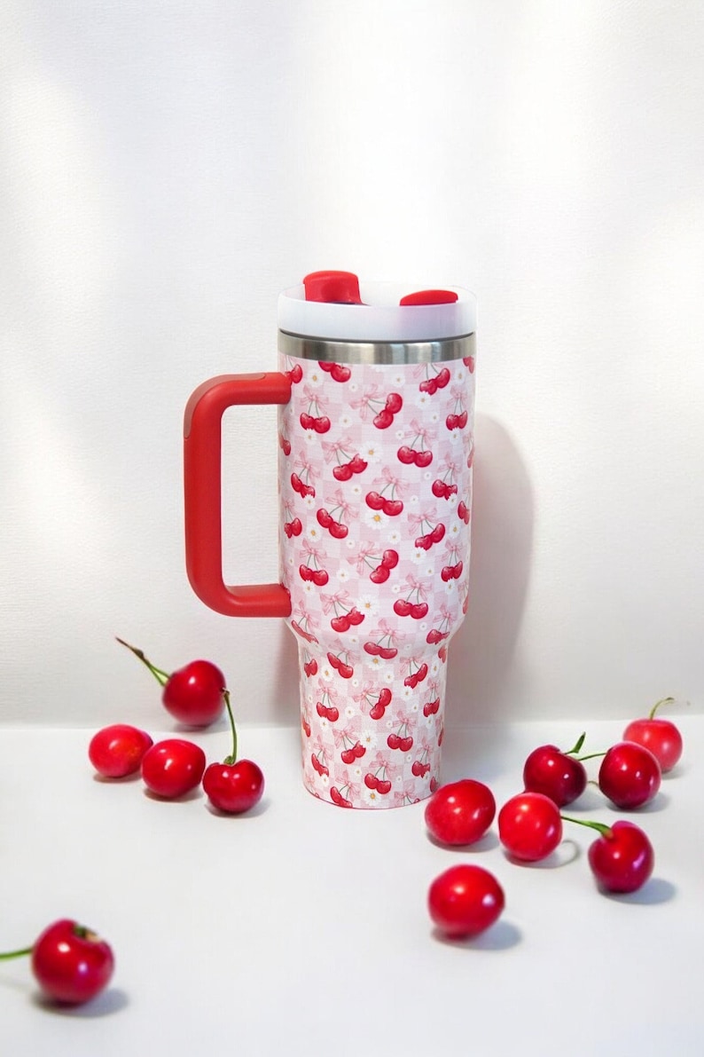 Puede incluir: Un vaso de acero inoxidable con asa y tapa rojas, decorado con un estampado de cerezas rojas y flores blancas sobre un fondo rosa de cuadros. Hay cerezas frescas esparcidas alrededor del vaso.