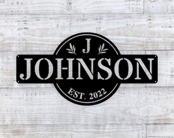 May include: Black metal sign with the last name "JOHNSON" in white lettering and a circle with the letter "J" and the text "EST. 2022" in white lettering.