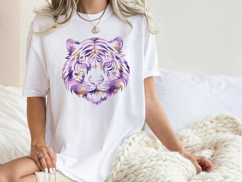 May include: A white t-shirt featuring a purple and gold tiger head graphic print.