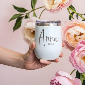 May include: A white insulated wine tumbler with a silver rim and lid. The name "Anna" and the word "Bride" are printed on the front in a dark gray script. The tumbler is held in front of a backdrop of pink and peach flowers.