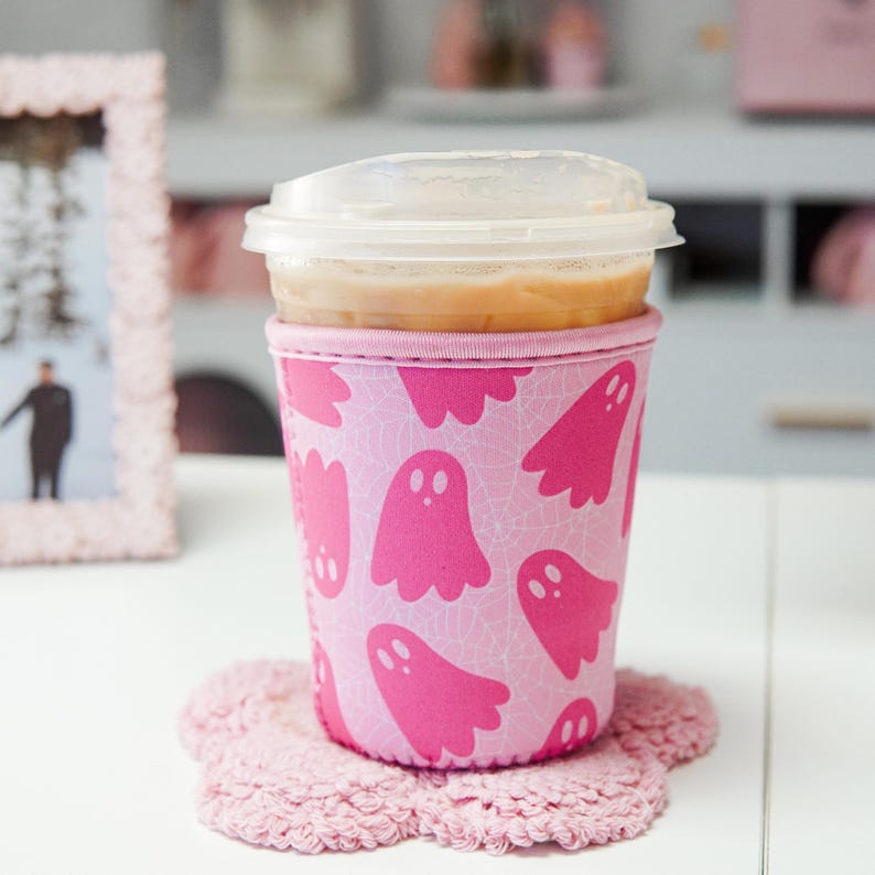 May include: A pink coffee cup sleeve featuring a ghost design, holding a clear plastic cup of iced coffee. The sleeve rests on a pink, flower-shaped coaster. The background is blurred, with a pink picture frame visible.